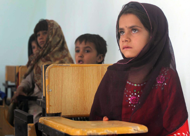 Eine Gruppe von Kindern sitzt an Tischen in einem Klassenzimmer in Afghanistan und nimmt an einem Unterricht teil, wobei eine Wand im Hintergrund zu sehen ist.