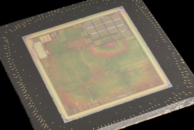 Nahaufnahme eines rechteckigen Halbleiterchips mit metallischem Glanz, umgeben von einem dünnen Metallrahmen und mit kleinen Löchern an seinen leicht erhöhten Kanten auf schwarzem Hintergrund.