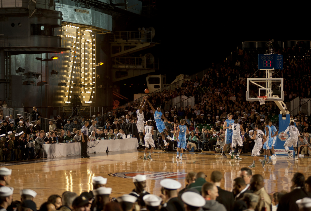 Eine Gruppe von Menschen, die Basketball auf einem Court mit einem Netz auf der rechten Seite spielen, mit Stühlen im Hintergrund und Geländern und Lampen sichtbar.