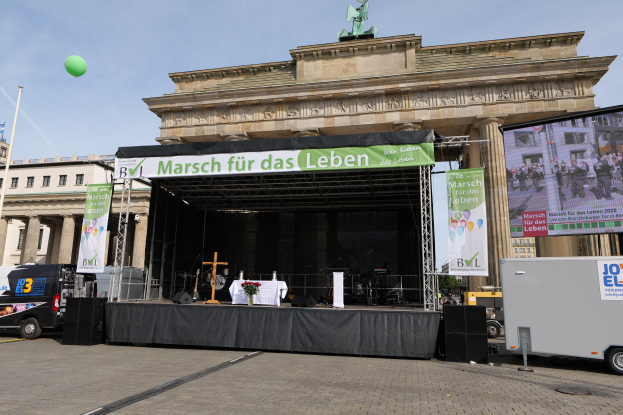 Bühne vor dem Brandenburger Tor mit einem Tisch, Bannern, Lautsprechern, Fahrzeugen, Gebäuden, einer Statue, einem Fahnenmast und einem bewölkten Himmel.