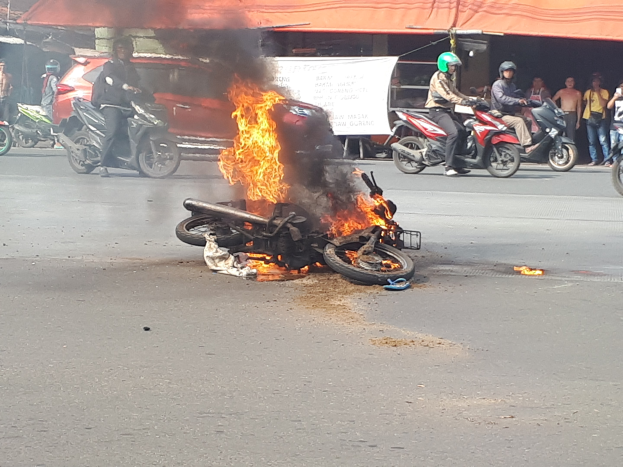 Ein in Flammen stehendes Motorrad am Straßenrand mit einem Auto und einem Van im Hintergrund, umgeben von Menschen, einige tragen Helme, eine Fahne mit Text und ein Zelt.