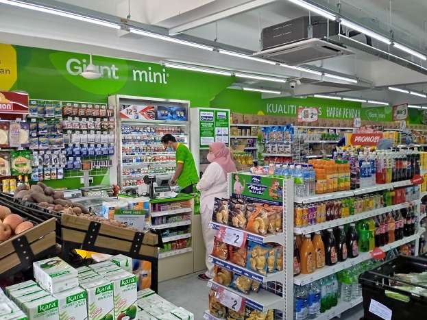 A brightly lit mini supermarket aisle stocked with bottles, boxes, fruits, and other food items on shelves, featuring ceiling-mounted boards, lights, and a projector.