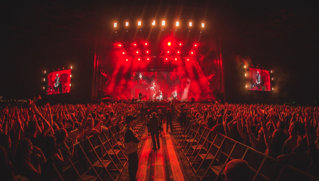 Eine große Menschenmenge steht vor einer hell erleuchteten Bühne mit Geländern, auf der Musiker spielen und andere zusehen, vor einem dunklen Hintergrund.