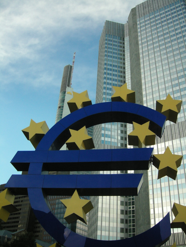 Euro sign in front of the European Central Bank headquarters in Frankfurt, Germany, with trees and a cloudy sky.