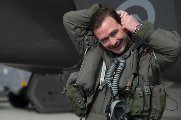 A smiling man in a fighter jet cockpit, wearing a jacket and watch, with hands on his head, in front of an aircraft in the background.