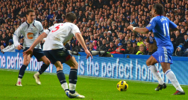 Personen mit einem Ball in der Vordergrund, eine Tafel mit Text in der Mitte, Kameras hinter der Tafel und eine Gruppe von Personen im Hintergrund.