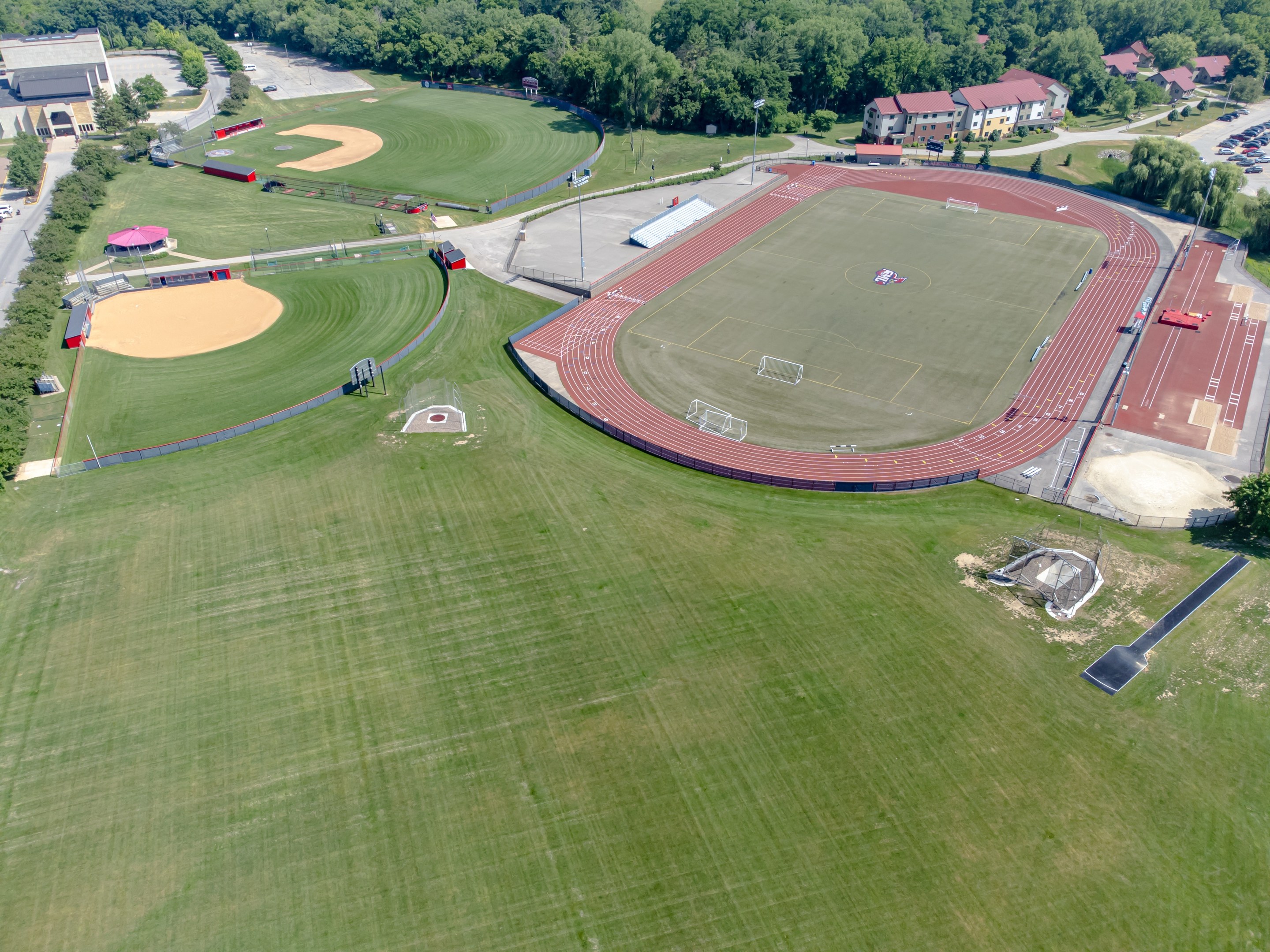 Aerial view of a baseball field with green grass, trees, buildings, and vehicles on a nearby road.