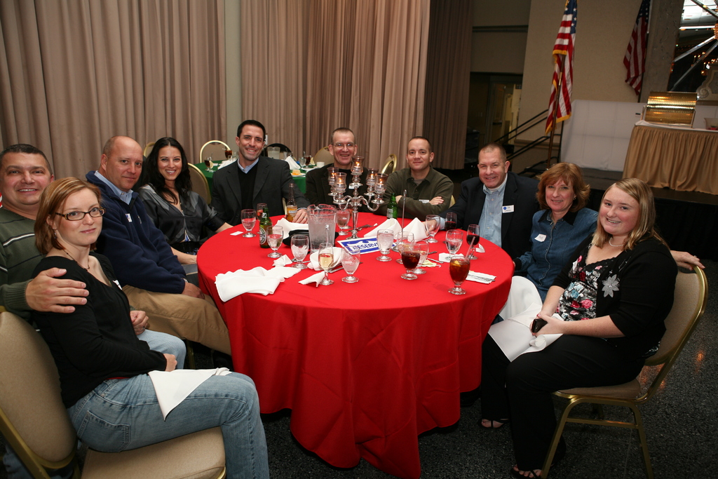 Eine Gruppe strahlender Menschen, die an einem Tisch mit Gläsern, Papieren, Weinflaschen und -gläsern, einer Flagge auf der rechten Seite und Vorhängen an der Wand sitzen.