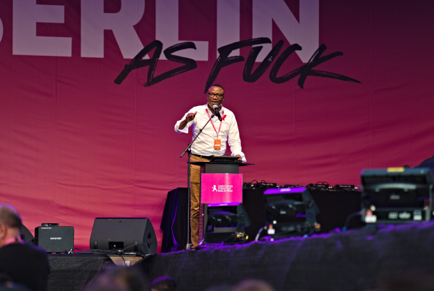 Mann in weißem Hemd und braunen Hosen, der in ein Mikrofon spricht, vor einem pinken 'Berlin as Fuck'-Plakat auf der Bühne