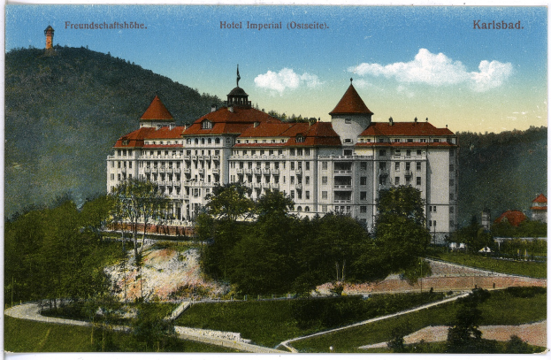 Eine alte Postkarte zeigt das Hotel Imperial in Karlsbad, Deutschland, ein großes Gebäude mit zahlreichen Fenstern, umgeben von Bäumen, Pflanzen und Gras, mit einem Weg im Vordergrund und Hügeln und einem bewölkten Himmel im Hintergrund, und Text oben auf dem Bild.