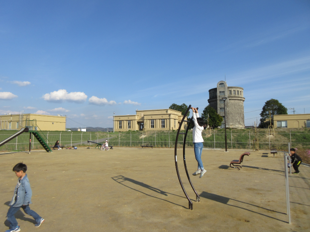 Eine Gruppe von Kindern spielt auf einem Spielplatz vor einem Gebäude mit Fenstern, umgeben von einem Metallzaun, Bänken, einer Treppe, einem Metallpfahl, einigen Pflanzen mit Blumen, Gras, der Rinde eines Baumes, einigen Pfählen, einem Turm, einigen Bäumen und einem bewölkten Himmel.