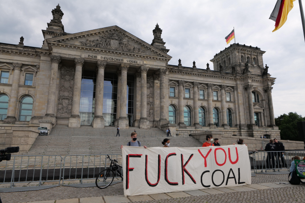 Gruppe von Menschen mit einem "Fuck You Coal"-Schild vor dem Reichstag in Berlin, Deutschland, mit seinen architektonischen Details, einem Fahrrad, Bäumen, einem Fahnenmast und einem bewölkten Himmel.