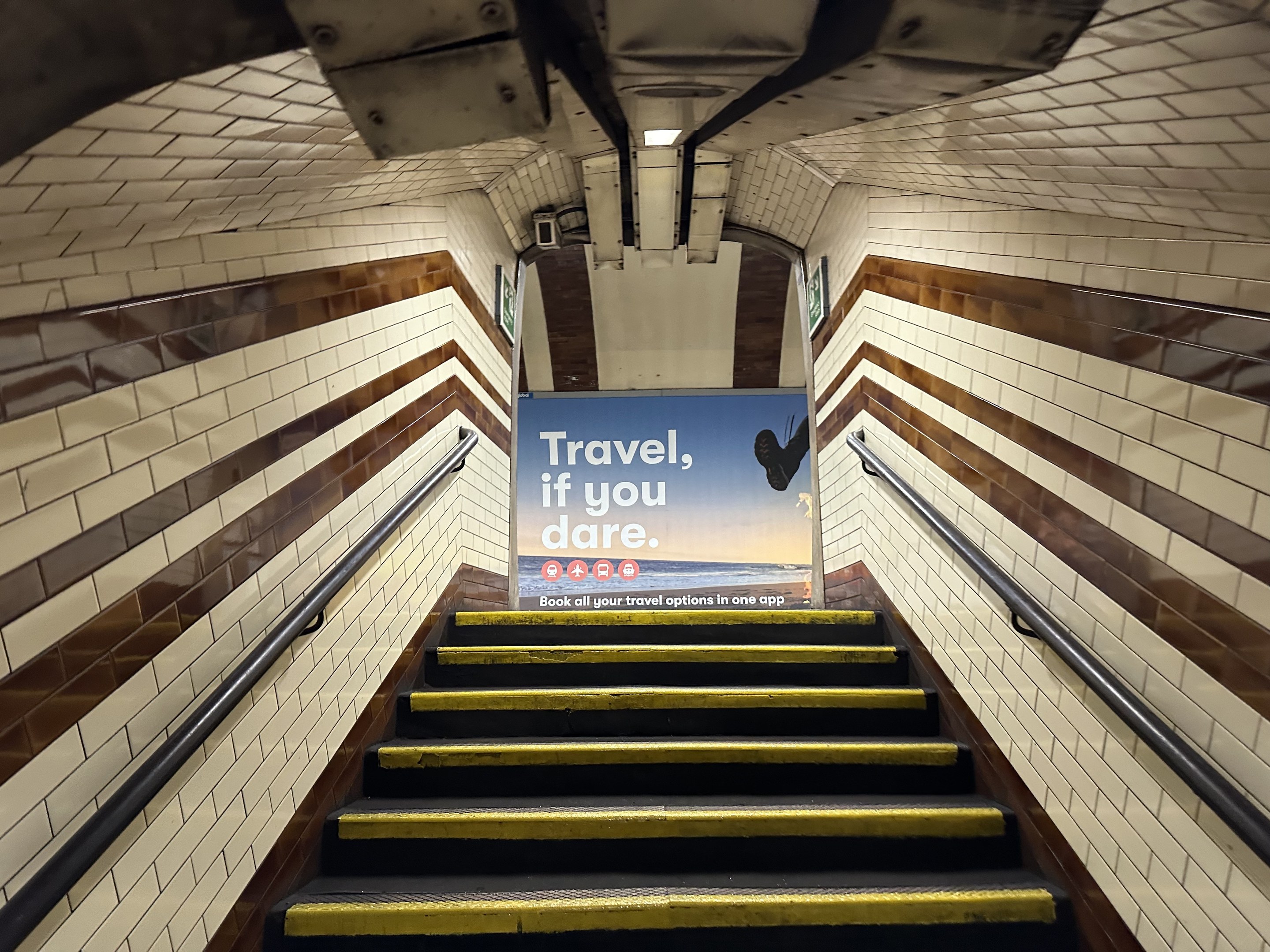 Eine Treppe in einer U-Bahn-Station mit einem Schild oben, das "Reise, wenn du wagst" trägt, flankiert von Geländern und sichtbaren Stationwänden.