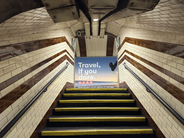 Eine Treppe in einer U-Bahn-Station mit einem Schild oben, das "Reise, wenn du wagst" trägt, flankiert von Geländern und sichtbaren Stationwänden.