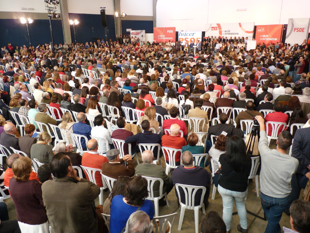 Ein volles Stadion mit vielen sitzenden und stehenden Menschen, eine Bühne mit zwei Rednern am Podium und mehreren Bannern und Lichtern in der Nähe.