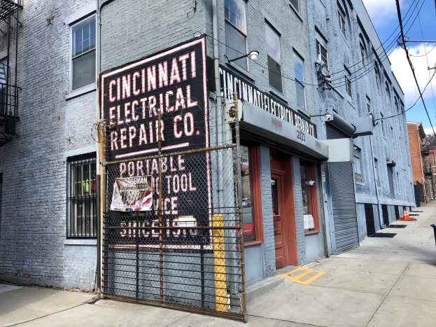 Außenansicht des Cincinnati Electrical Repair Co.-Gebäudes mit einer Schautafel, einem Metallzaun, einem Müllbehälter, Pfählen, Drähten, Bäumen und einem bewölkten Himmel.