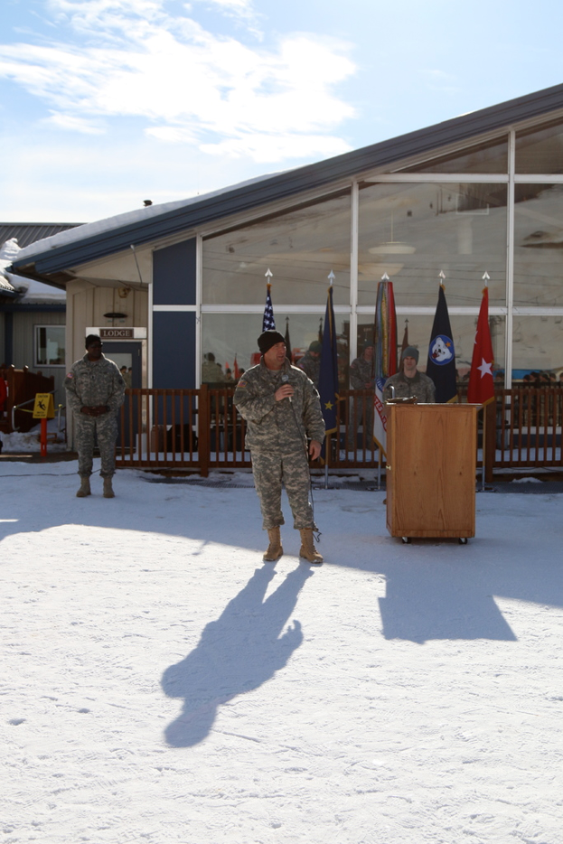 Eine Gruppe von Menschen steht in der Nähe eines Podiums mit Flaggen, einem Zaun und Hütten im Hintergrund unter einem bewölkten Himmel.