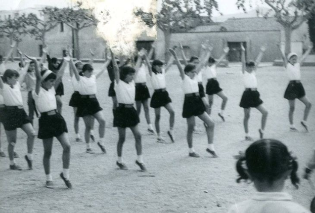Schwarzes und weißes Bild von jungen Mädchen in Schuluniformen, die auf einer Straße tanzen, die von Gebäuden und Bäumen gesäumt ist.