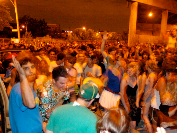 Menschen tanzen vorne vor einem Konzert mit Lichtern, Polen, Bäumen und anderen Gegenständen im Hintergrund.