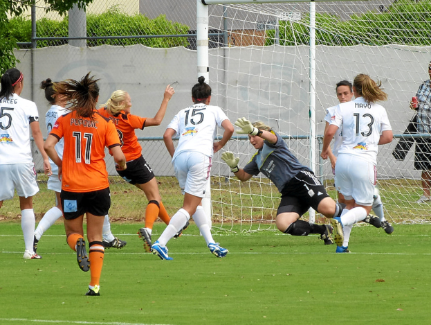 Eine Gruppe von Frauen spielt ein Spiel im Freien mit einem Maschendrahtzaun und Bäumen im Hintergrund.