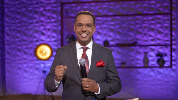 Stephen Colbert, Moderator von *The Daily Show with Trevor Noah*, steht auf einer Bühne und hält ein Mikrofon in der Hand, trägt einen Anzug, eine Krawatte und eine Brille, während hinter ihm Lichtausrüstung und Gegenstände zu sehen sind.