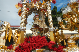 Ein Rosenmontagszug-Schmuckwagen mit roten Rosen und goldenen Verzierungen, darunter Statuen und Blumen, vor einem Gebäude, Bäumen und einem bewölkten Himmel.