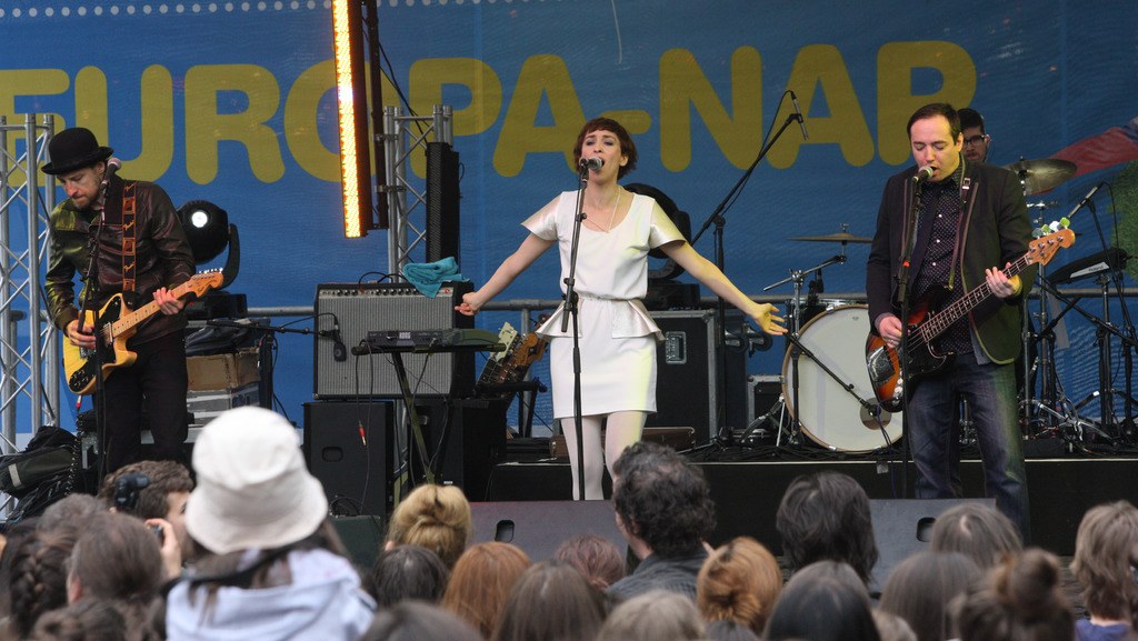 Vier Musiker auf der Bühne: Eine Frau in einem weißen Kleid singt in der Mitte, flankiert von zwei Männern, die Gitarren spielen, mit einem Publikum unten und Lautsprechern an der Wand im Hintergrund.