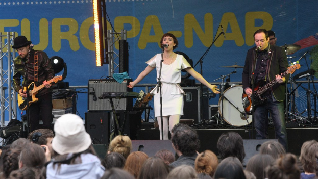 Vier Musiker auf der Bühne: Eine Frau in einem weißen Kleid singt in der Mitte, flankiert von zwei Männern, die Gitarren spielen, mit einem Publikum unten und Lautsprechern an der Wand im Hintergrund.