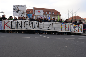 Demonstranten mit einem Transparent mit der Aufschrift "Kein Grund zur Feier" gegen deutsche Sparmaßnahmen, im Hintergrund Straßeninfrastruktur und Gebäude.