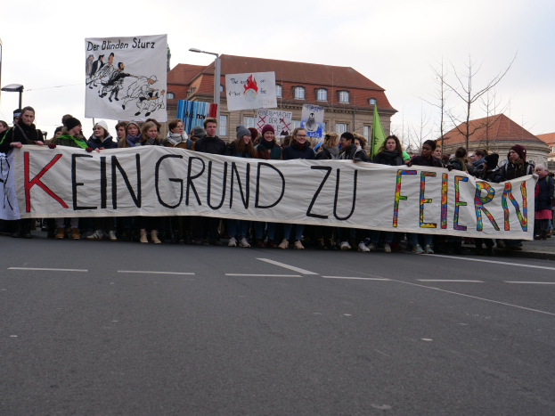 Demonstranten mit einem Transparent mit der Aufschrift "Kein Grund zur Feier" gegen deutsche Sparmaßnahmen, im Hintergrund Straßeninfrastruktur und Gebäude.