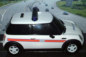 White police car on a road with multiple vehicles, grass and trees on either side, outside a city.