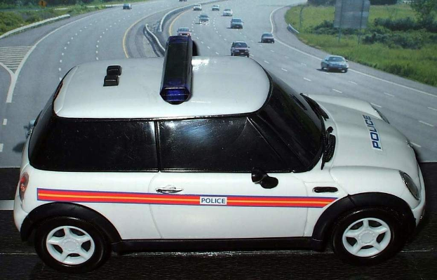 White police car on a road with multiple vehicles, grass and trees on either side, outside a city.
