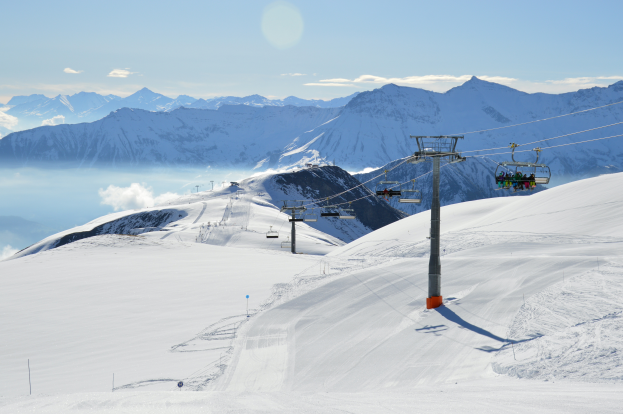 Eine Sesselbahn fährt einen schneebedeckten Berg hoch, auf der Menschen auf Sesseln sitzen, die an Stangen und Seilen hängen, mit schneebedeckten Hügeln und einem bewölkten Himmel im Hintergrund.