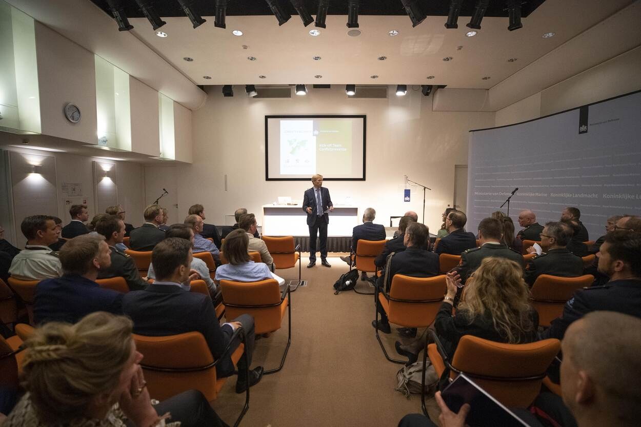 A man standing and speaking into a microphone while a group of people sit in chairs facing a projector screen displaying text, with a clock and ceiling lights visible in the background.