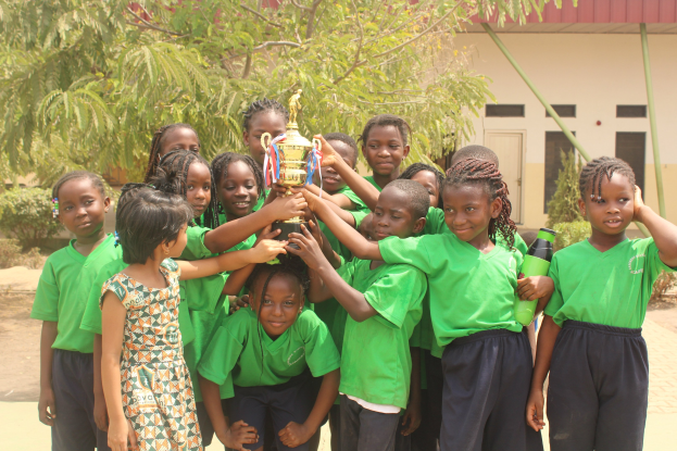 Eine Gruppe von Kindern hält einen Pokal in einer Schulumgebung mit Bäumen und einem Gebäude im Hintergrund.