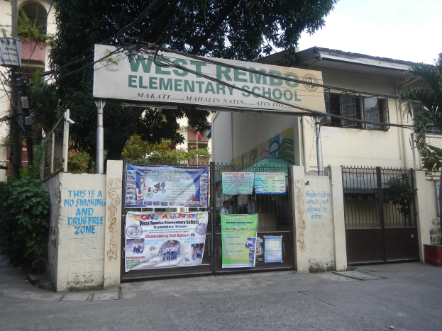 Außenansicht der West Rembo-Grundschule in Manila, Philippinen, die ein Gebäude mit Fenstern, ein Tor, eine Tafel mit Text, Plakate an der Wand, Pflanzen, Bäume, einen Straßenmast, Drähte und einen bewölkten Himmel zeigt.