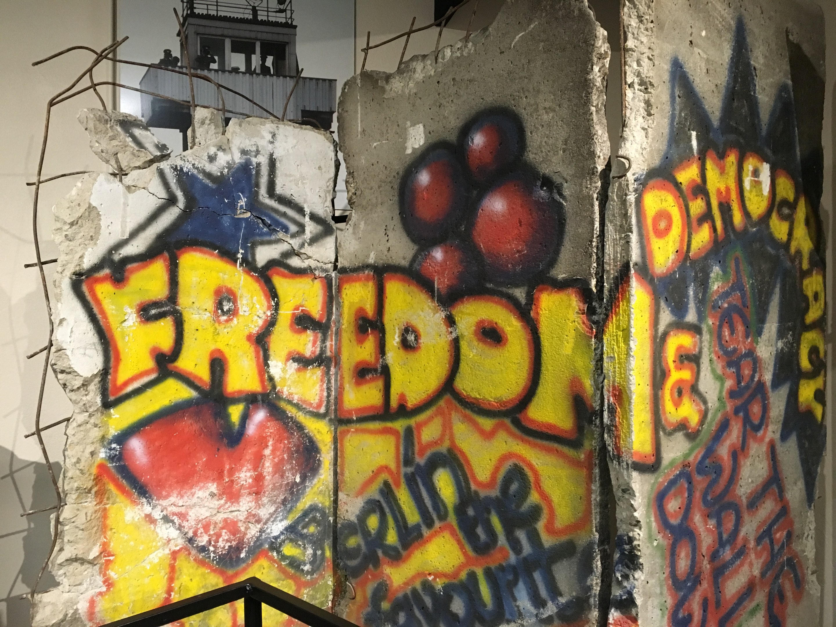 Mauer mit Graffiti, das in großen schwarzen Buchstaben "Freiheit und Demokratie" liest, eine Reling unten und ein Gebäude mit Fenstern im Hintergrund.