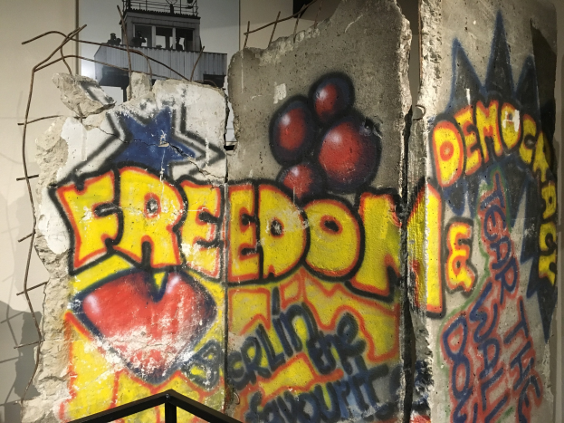 Mauer mit Graffiti, das in großen schwarzen Buchstaben "Freiheit und Demokratie" liest, eine Reling unten und ein Gebäude mit Fenstern im Hintergrund.