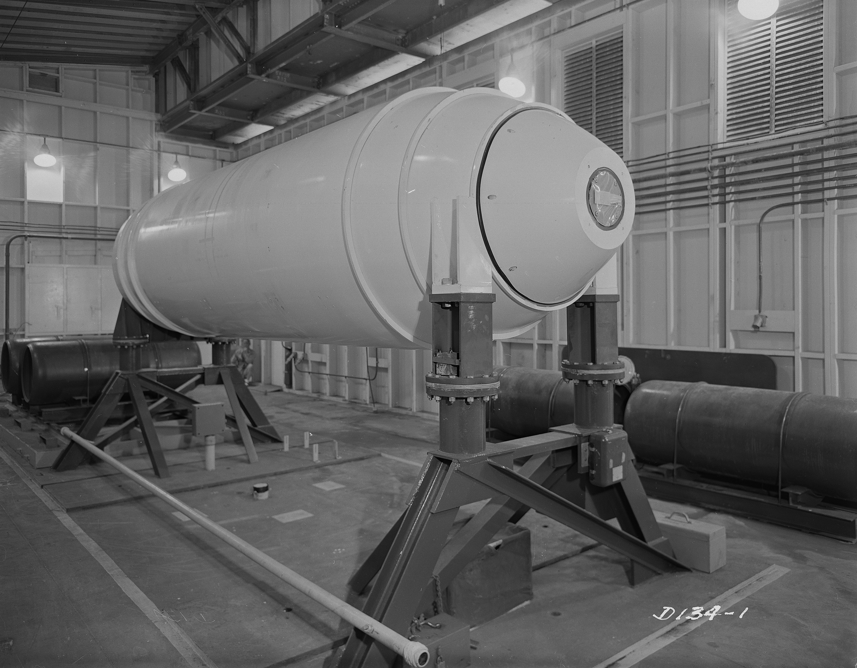 Schwarzes und weißes Foto einer großen zylindrischen Wasserstoffbombe auf einem Ständer in einer Fabrik, mit Rohren, Lampen und einer Wand im Hintergrund und Text unten.