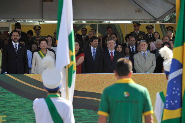 Menschen vor Fahnen mit einem Banner im Hintergrund und mehr Menschen unter einem Zelt.