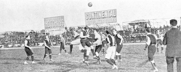 Ein Schwarz-Weiß-Bild von einer Gruppe von Männern, die auf einem Feld Fußball spielen, mit einem Ball in der Luft und Zuschauern im Hintergrund, mit Tafeln, die Text über das Spiel anzeigen.