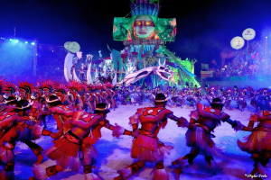 Eine Gruppe von Menschen in farbenfrohen Kostümen tanzt auf einer Bühne vor einem sitzenden Publikum, beleuchtet von Scheinwerfern und umgeben von Pfählen und Ballons, mit einem Bildschirm, der eine Person und andere Gegenstände im Hintergrund zeigt, während des Karnevals 2014 in Rio.