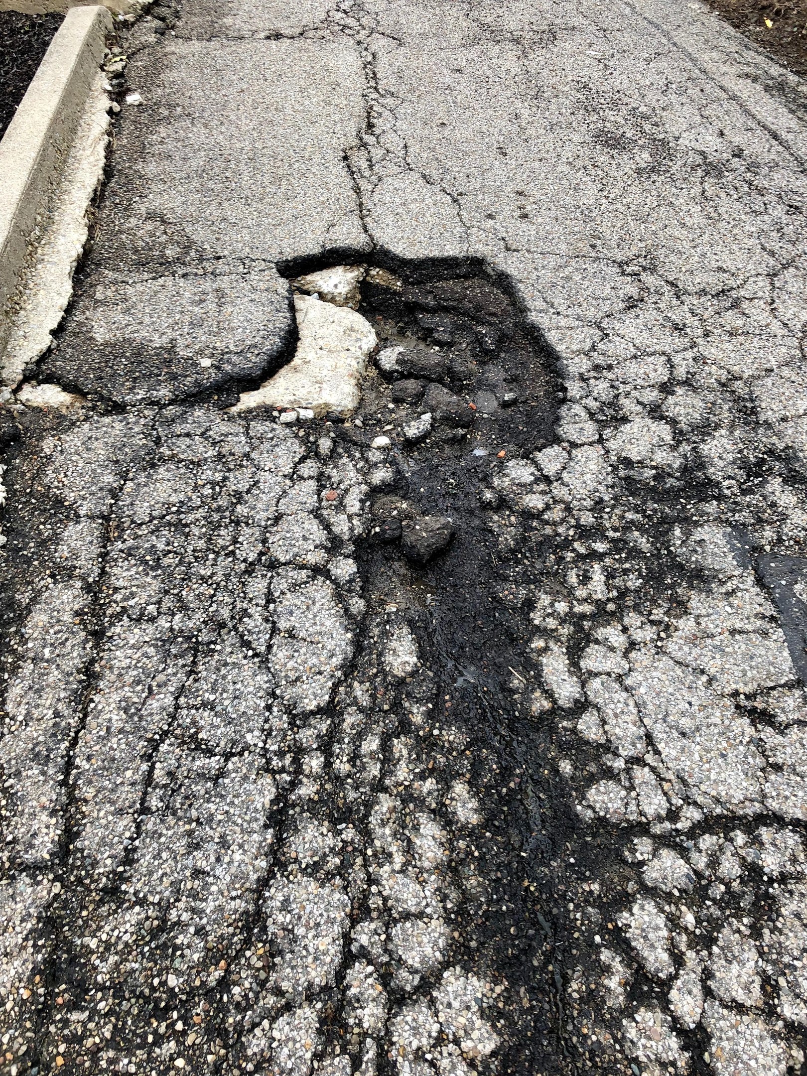 Eine Straße mit einem großen Schlagloch in der Mitte, verstreute kleine Steine und eine Wand auf der linken Seite.