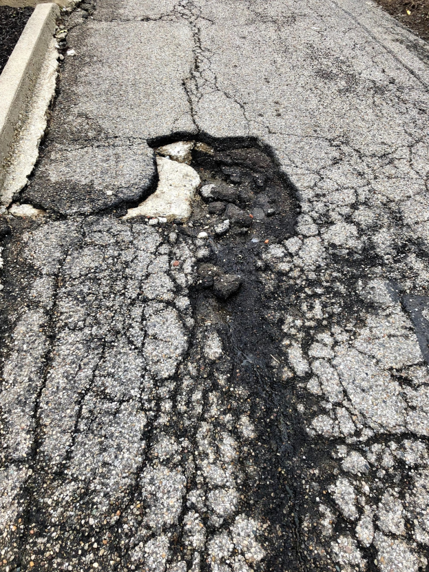 Eine Straße mit einem großen Schlagloch in der Mitte, verstreute kleine Steine und eine Wand auf der linken Seite.
