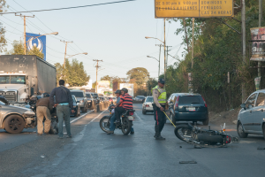 Eine Gruppe von Menschen steht um ein verunglðcktes Motorrad auf der Seite einer Straße mit mehreren Fahrzeugen, darunter ein Lastwagen, und einer Hintergrund von Bäumen, Masten, Lichtern und Schildern.