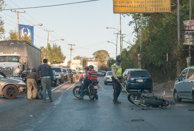 Eine Gruppe von Menschen steht um ein verunglðcktes Motorrad auf der Seite einer Straße mit mehreren Fahrzeugen, darunter ein Lastwagen, und einer Hintergrund von Bäumen, Masten, Lichtern und Schildern.