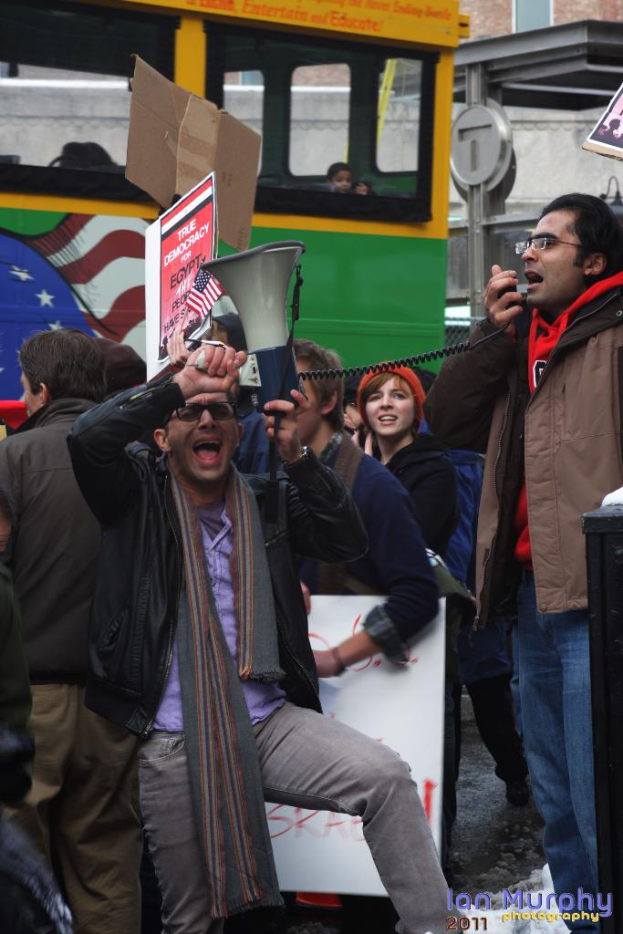 Eine Gruppe von Menschen mit Schildern, Lautsprechern und Mikrofonen vor einem Bus und Gebäuden.