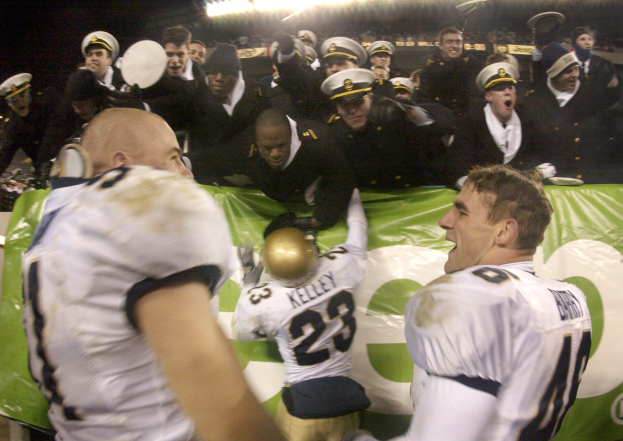 Eine Gruppe von Männern in Mützen steht auf einem Fußballfeld und hält ein Banner, wobei einer der Männer einen Helm hält und Scheinwerfer das Feld beleuchten.