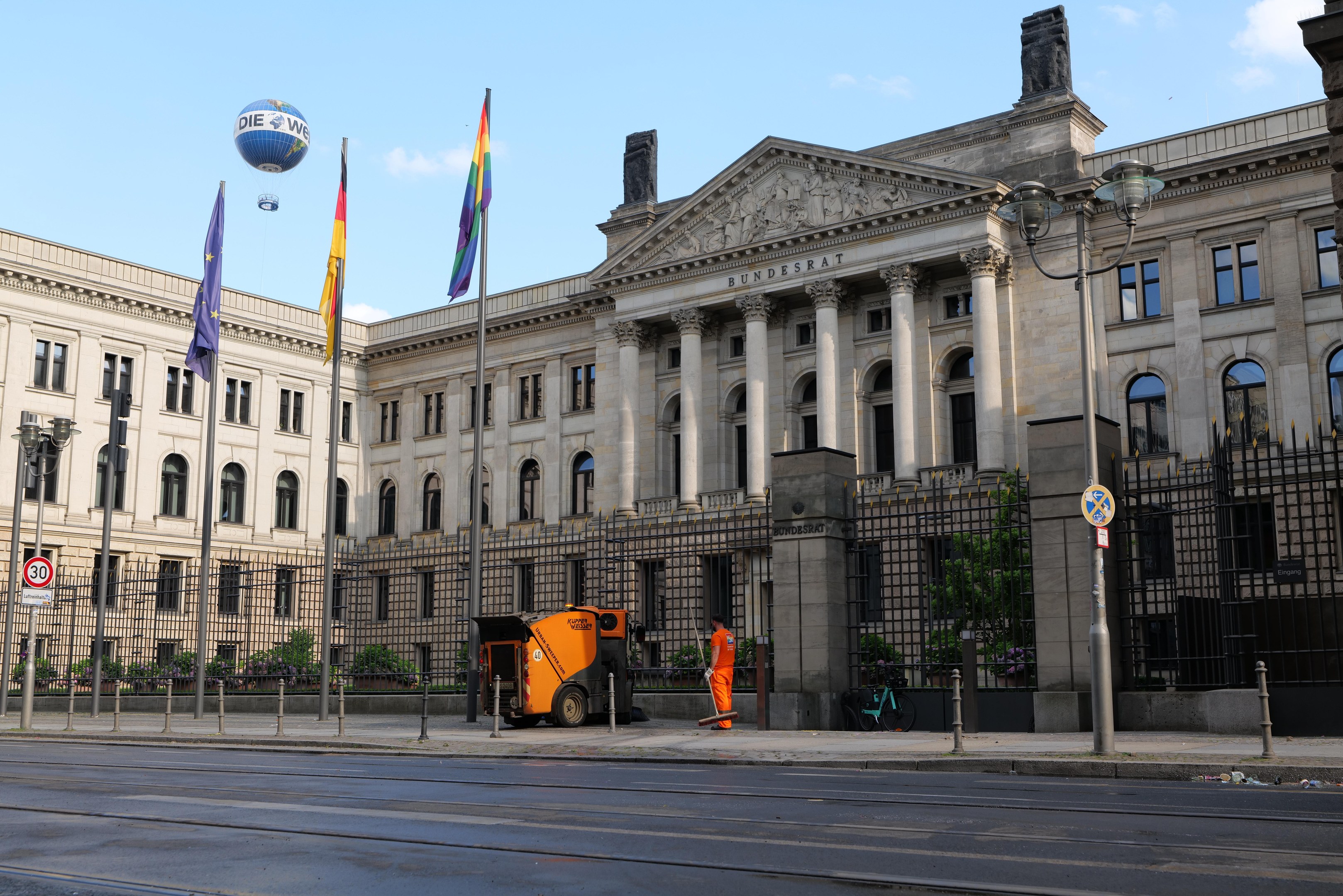 Large modern building with many windows, identified as the Bundestag in Berlin, Germany, surrounded by street infrastructure, pedestrians, trees, and a cloudy sky.