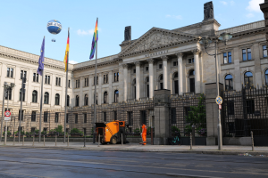 Large modern building with many windows, identified as the Bundestag in Berlin, Germany, surrounded by street infrastructure, pedestrians, trees, and a cloudy sky.